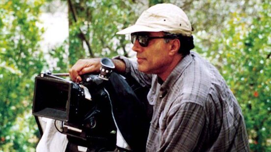 Abbas Kiarostami, wearing glasses and a cap, directs a movie with his camera by his side.