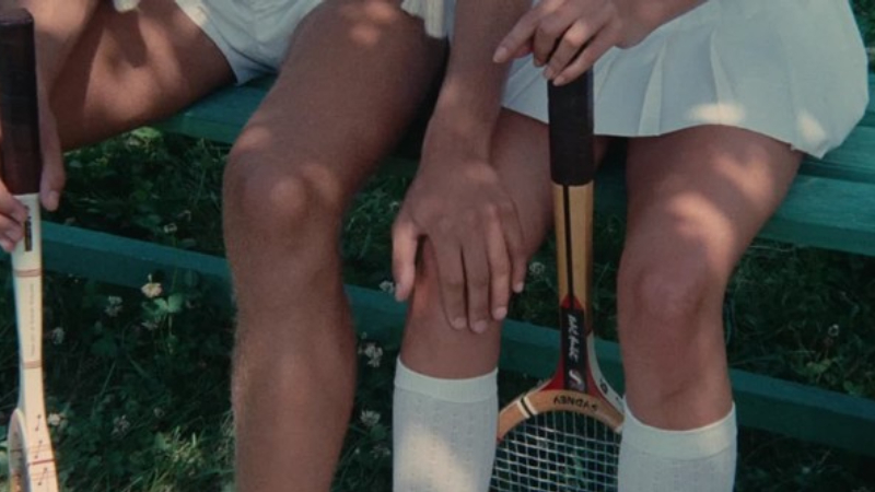 In the film "Claire's Knee," a young boy with a tennis racket touches the knee of a teenage girl as they sit on a bench, depicted from below the waist.
