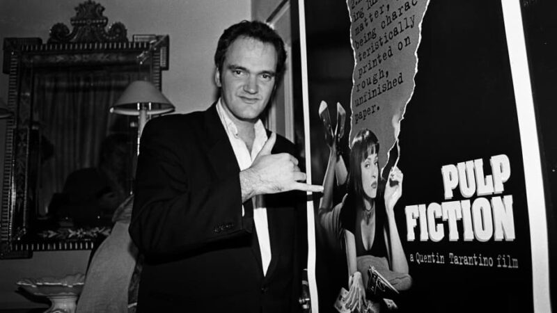 Quentin Tarantino standing beside a Pulp Fiction movie poster.
