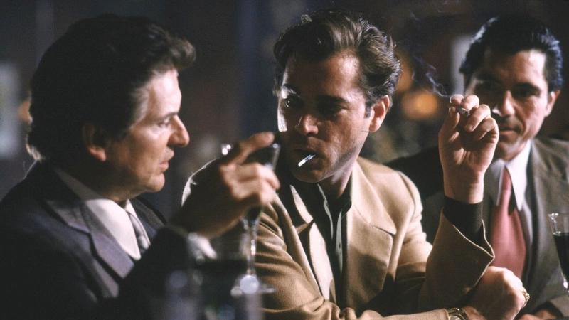 Ray Liotta as Henry Hill engages in conversation with Joe Pesci as Tommy DeVito, holding a cigarette and a spoon in his mouth. In the background, Joseph Bono as Mikey Franzese observes them.