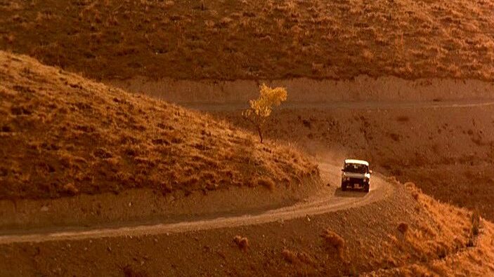 Homayoun Ershadi as Mr. Badii in Taste of Cherry, his car depicted from a distance in a desert environment. Minimalist shot.