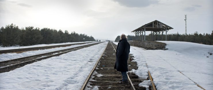 Haluk Bilginer as Aydin from the movie Winter Sleep, standing on a snowy train track, hands in coat pockets, gazing into the distance.