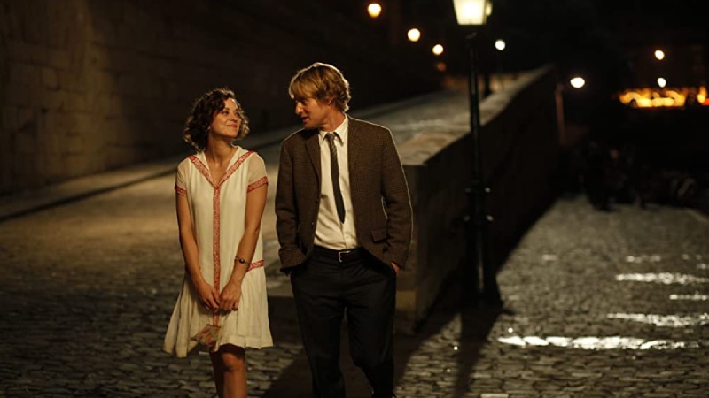 Two characters from the movie "Midnight in Paris," Gil and Adriana, walk together in a dimly lit environment at night, looking at each other.
