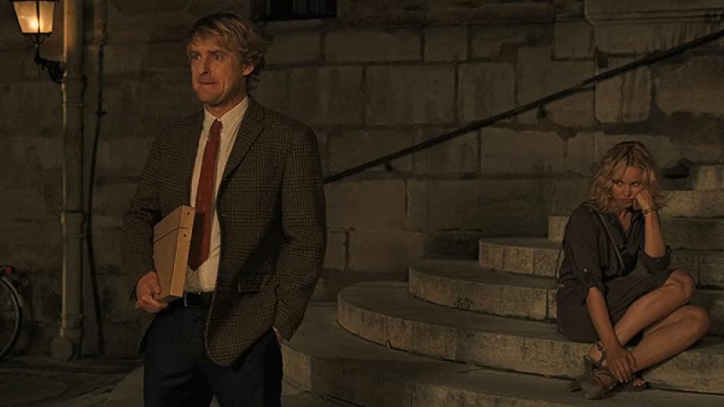 Gil, a character from the movie "Midnight in Paris," stands with his back to Inez, looking contemplative in a nighttime Parisian setting. Inez is sitting on a flight of stairs nearby, looking tired and drained.