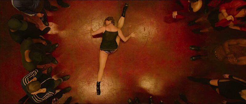 A girl from the movie Climax performs a provocative dance with her legs apart while the surrounding dancers watch.