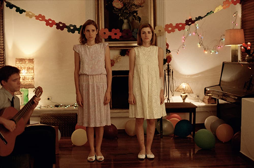 A still image from the Greek film "Dogtooth" showing two sisters standing side by side in a dimly lit room, both dressed in identical pink dresses and staring blankly ahead.