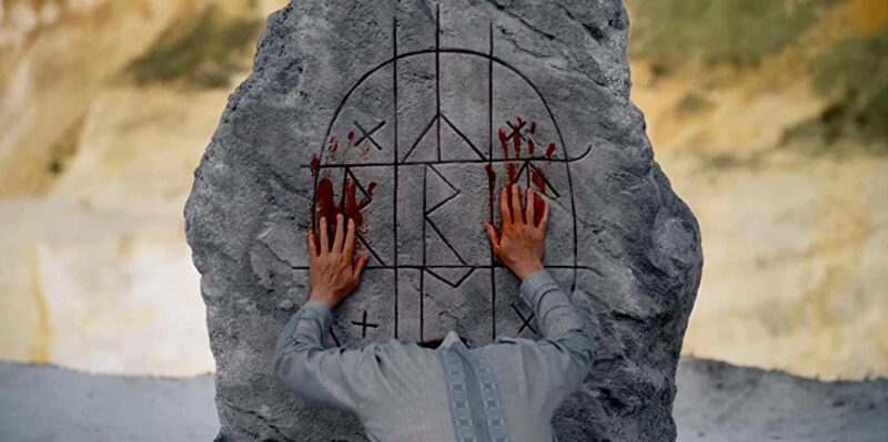 Man with bloody hands performing ritual on rock with symbolic drawings