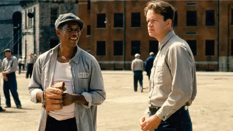 Tim Robbins as Andy Dufresne from The Shawshank Redemption, deep in thought. Morgan Freeman as Ellis Boyd 'Red' Redding, holding a baseball glove, smiles while looking at Andy.