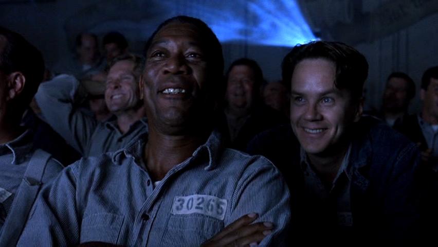 A still from the movie "The Shawshank Redemption," depicting a group of inmates gathered together in the prison yard, watching a movie projected onto a sheet.
