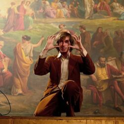 10 Signature Elements of Wes Anderson's Filmmaking Style! A photograph of Wes Anderson on a film set, crouched down and directing an actor in a specific pose.