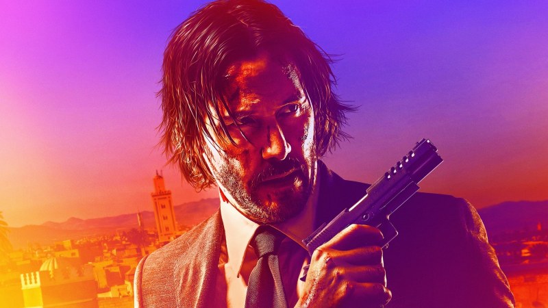 John Wick, a man with short black hair, wearing a black suit and holding a gun, looks to his right with a serious expression on his face.