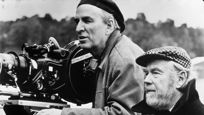 Ingmar Bergman stands behind a movie camera, giving instructions to actors on set.