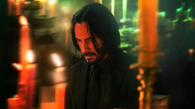 John Wick, a man with short black hair, wearing a black suit, looks down with a thoughtful expression on his face.