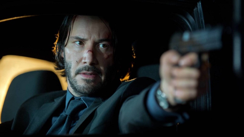 John Wick, a man with short black hair, wearing a black suit and holding a gun, points the weapon threateningly in front of him.