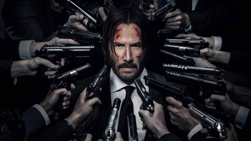 John Wick, a man with short black hair, wearing a black suit and holding a gun, surrounded by multiple armed men in a dimly-lit room.