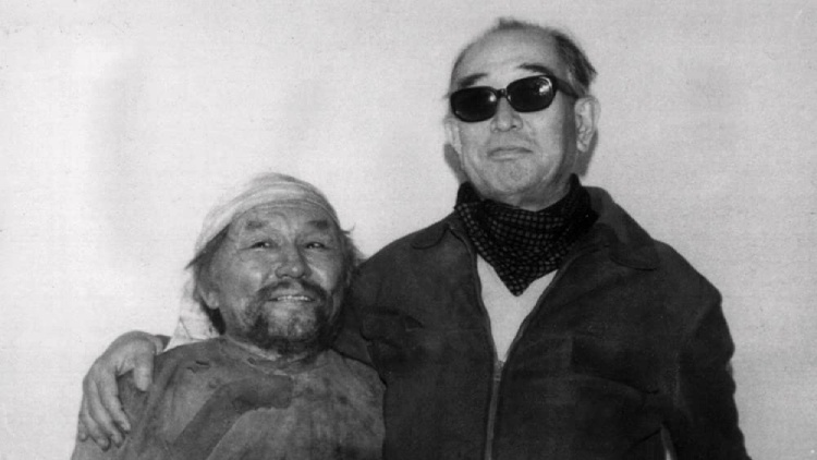 Akira Kurosawa, donning glasses, warmly smiles and places a reassuring hand on the shoulder of a samurai cast member next to him.