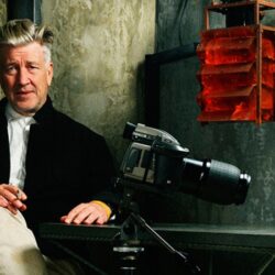 10 Signature Elements of David Lynch's Filmmaking Style! David Lynch, the enigmatic filmmaker, crosses his legs with a cigarette in hand, his trusty DSLR camera placed in front of him.
