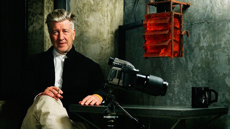 David Lynch, the enigmatic filmmaker, crosses his legs with a cigarette in hand, his trusty DSLR camera placed in front of him.