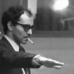 10 Signature Elements of Jean-Luc Godard's Filmmaking Style! Renowned filmmaker Jean-Luc Godard with a cigarette in his mouth, directing the film production team.