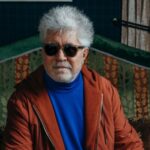 Pedro Almodóvar, wearing glasses, strikes a charismatic pose against a vibrant backdrop, his colorful attire complementing the scene.