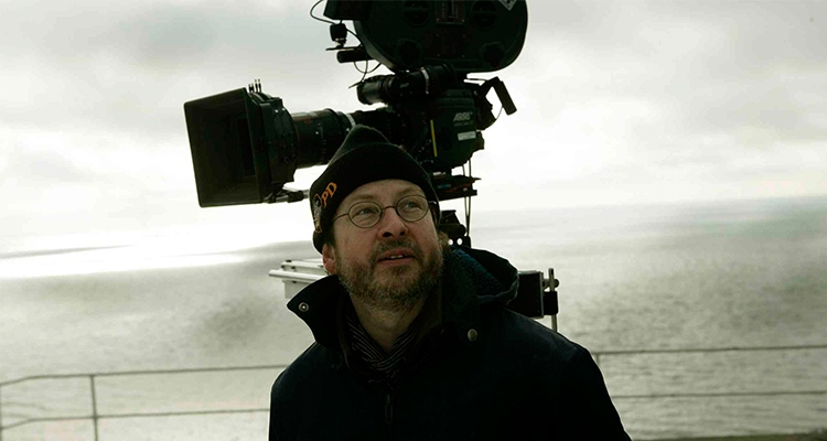 Lars von Trier wearing glasses, glancing upward to his left, sporting a subtle smile, with a camera visible in the background.