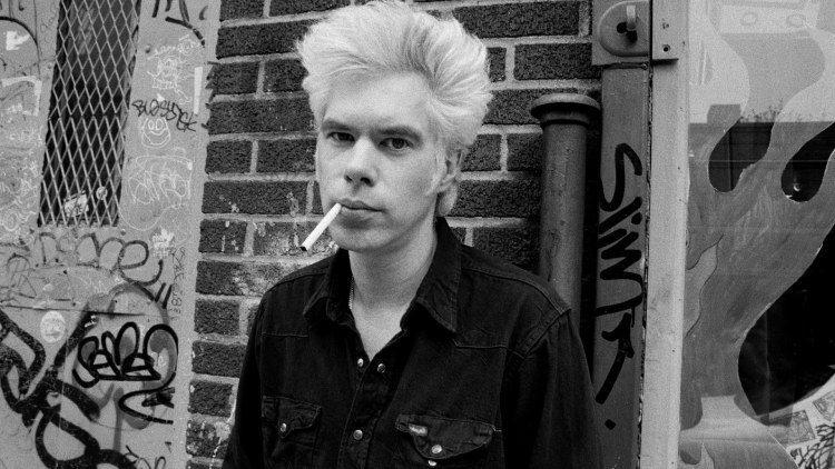 Iconic black and white photograph of Jim Jarmusch, donning a black shirt, with a cigarette poised in his mouth.
