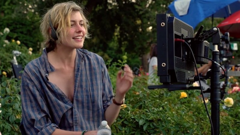 Greta Gerwig, with a subtle smile, directs a movie while wearing headphones, with an open shirt and a screen in front of her.