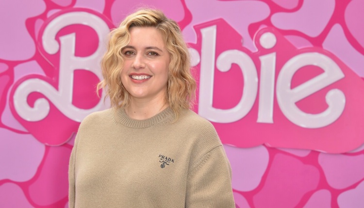 Greta Gerwig smiles with the 'Barbie' movie logo, which she directed, prominently displayed behind her.