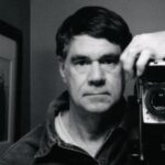 10 Signature Elements of Gus Van Sant's Filmmaking Style! Gus Van Sant looks directly at the camera, with his camera positioned closely beside him.