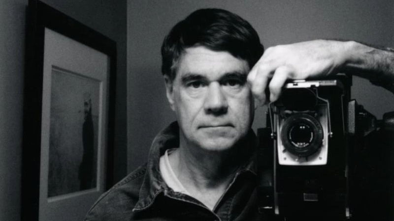 Gus Van Sant looks directly at the camera, with his camera positioned closely beside him.