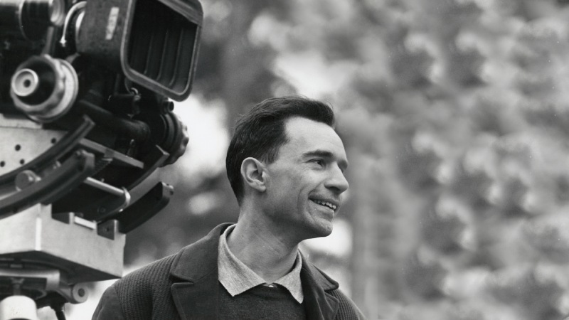 Director Jacques Rivette, with a beaming smile, stands behind a film camera, radiating joy and passion for filmmaking.