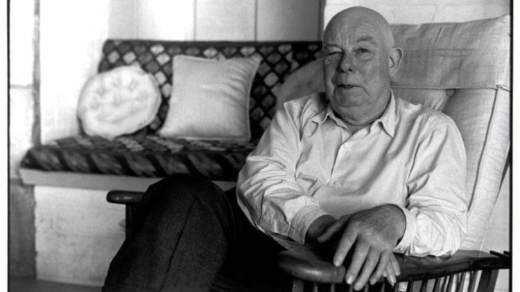 Jean Renoir strikes a pose while seated on a sofa, exuding an air of confidence.