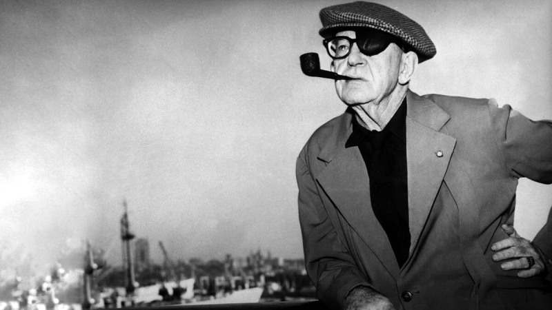 John Ford, one eye bandaged, holding a pipe in his mouth and resting one hand on his waist.