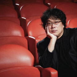 Director Bong Joon-ho seated in a movie theater, resting his chin on his hand, with a thoughtful expression, as he gazes at the camera.