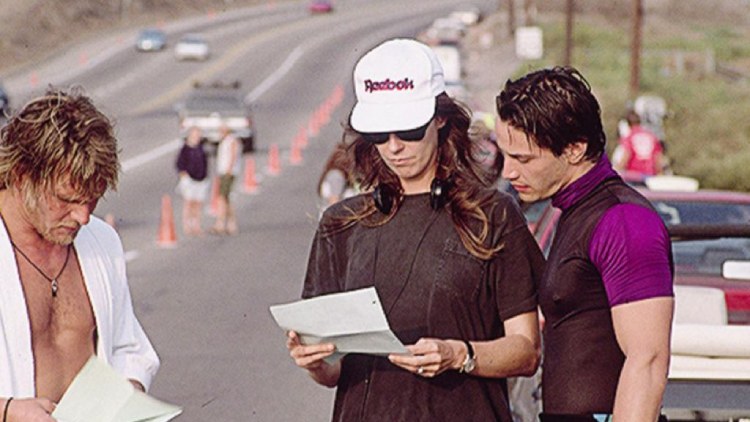 Kathryn Bigelow, script in hand and headphones in ear, collaborates with Keanu Reeves who seeks guidance on his role.