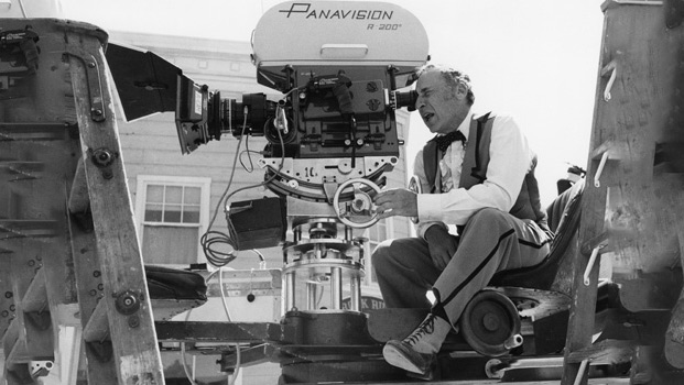 Mel Brooks, seated in the director's chair, peers through the viewfinder of his Panavision camera while actively directing a movie.