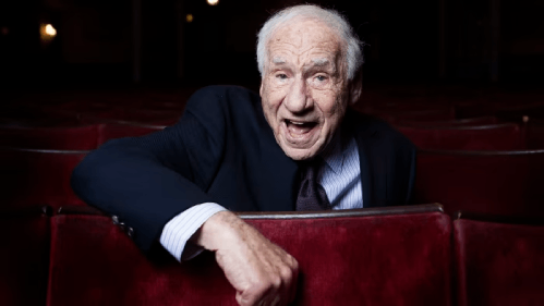 Mel Brooks, dressed in a suit, sits in a movie theater, exuding a joyful and humorous expression.