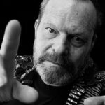10 Signature Elements of Terry Gilliam's Filmmaking Style! Terry Gilliam strikes a dynamic pose, gesturing towards the camera with expressive hand movements.