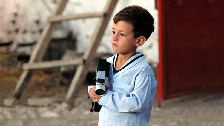 Ege Tanman, portraying Deniz in the movie "Babam ve Oğlum (My Father and My Son)" appears pensive in his pajamas, holding a handheld camera. Short Description: A reflective moment from "My Father and My Son," featuring Ege Tanman as Deniz, dressed in pajamas and holding a handheld camera, reflecting the film's narrative.