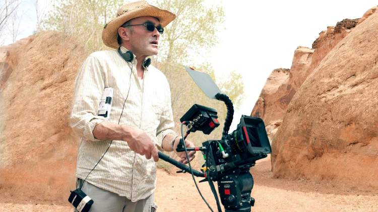 Danny Boyle, sporting a hat and sunglasses, holds a handheld camera as he directs a film.