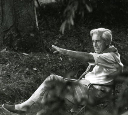 Director Robert Bresson sits in a garden and points at something with his hand.
