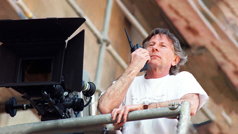 Director Roman Polanski, holding a walkie-talkie, stands next to a camera, actively directing a film.