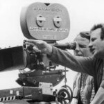 10 Signature Elements of Michael Mann’s Filmmaking Style! Michael Mann beside a camera on a tripod, overseeing the director of photography's work while directing the film with hand gestures.