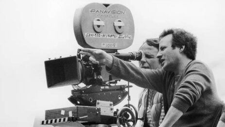 Michael Mann beside a camera on a tripod, overseeing the director of photography's work while directing the film with hand gestures.