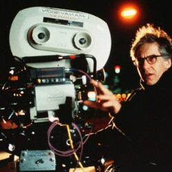 10 Signature Elements of David Cronenberg’s Filmmaking Style Image of director David Cronenberg on a movie set, gesturing with his hands while directing a scene with a camera in the foreground.
