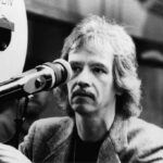 10 Signature Elements of John Carpenter’s Filmmaking Style! John Carpenter on a film set, peering through a camera viewfinder while directing a scene.