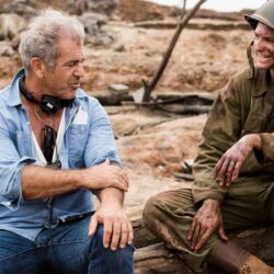 10 Signature Elements of Mel Gibson's Filmmaking Style! Director Mel Gibson on the set of the movie "Hacksaw Ridge" with headphones in his neck. Seated next to him is an actor from the film, engaged in conversation and sharing laughter.