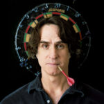 10 Signature Elements of Jay Roach's Filmmaking Style! Director Jay Roach humorously poses in front of a target board, with a dart's arrow placed playfully in his mouth.