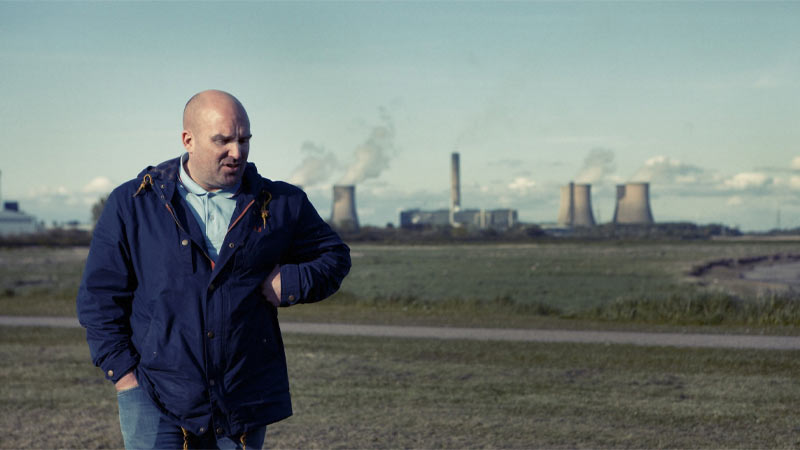 Director Shane Meadows strolls with a hand in his pocket, framed by smoking factories in the background.
