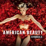 What "American Beauty" Symbols Really Mean? — Plastic Bag, Roses, and More! Mena Suvari as Angela Hayes lying among roses in the iconic bird's eye view shot from the movie American Beauty. Text overlay reads 'American Beauty symbols.